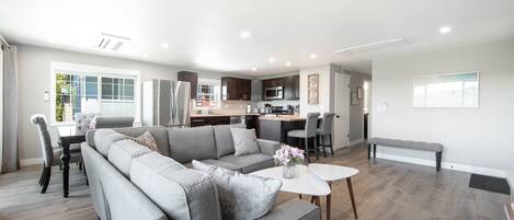 A modern living room with a grey sectional sofa, a dining table, and a kitchen area in the background.