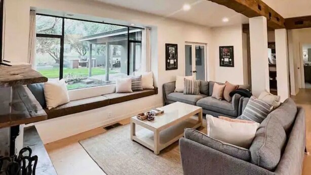A modern living room with a large window, a sofa set, a coffee table, and a fireplace. - Luxurious 3-BR Home: BBQ Grill, Hot Tub & Fire Pit (Angels Camp)