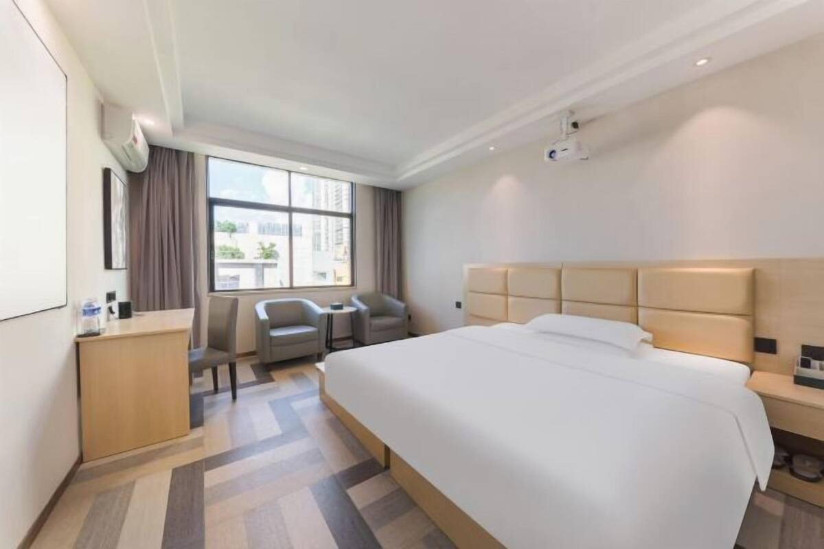 Habitación de hotel con una cama grande, un escritorio, dos sillas, una ventana con cortinas y un proyector en el techo.