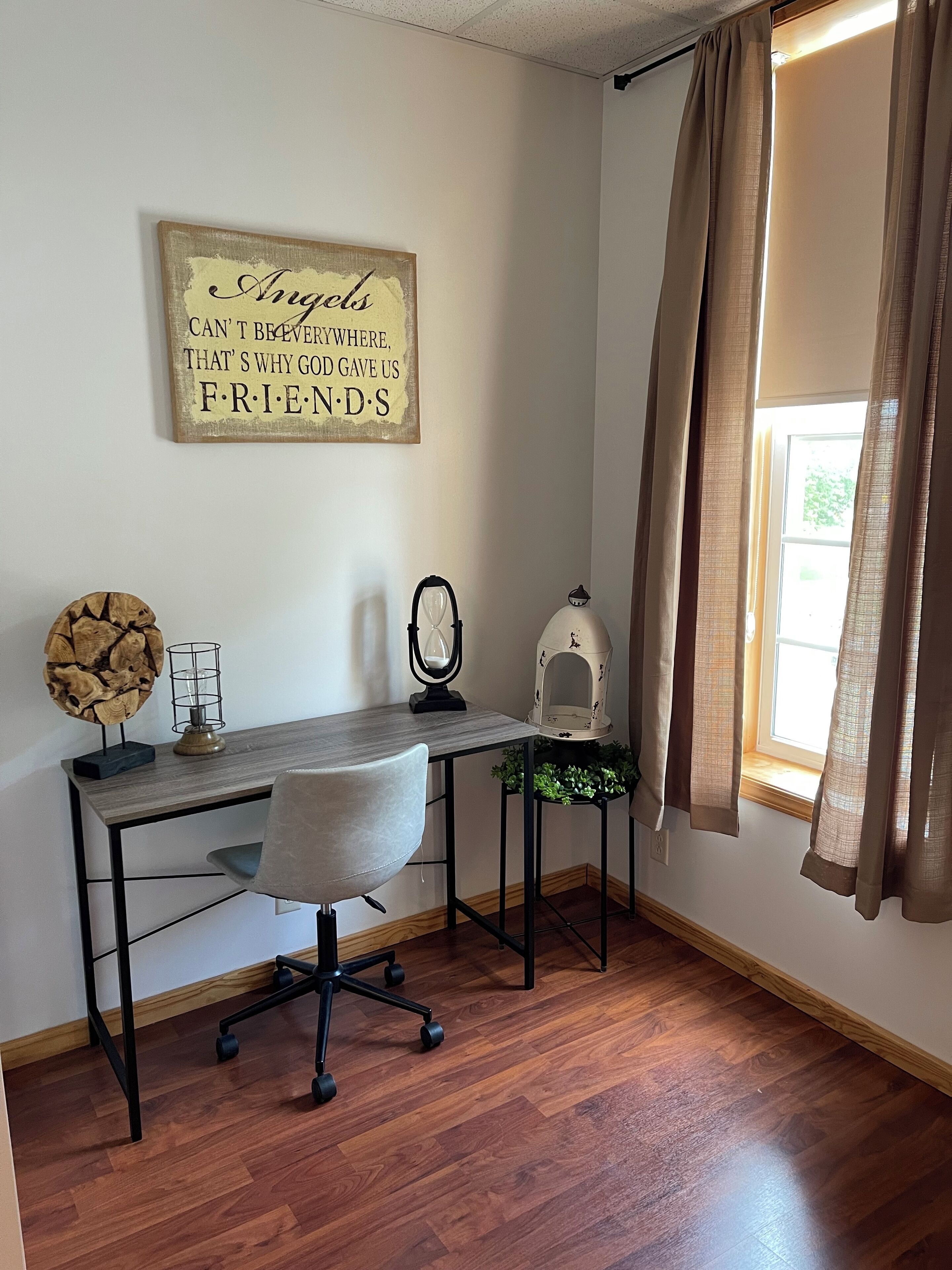 A room with a wooden desk, a chair, a wall clock, a small plant, and a window with curtains.