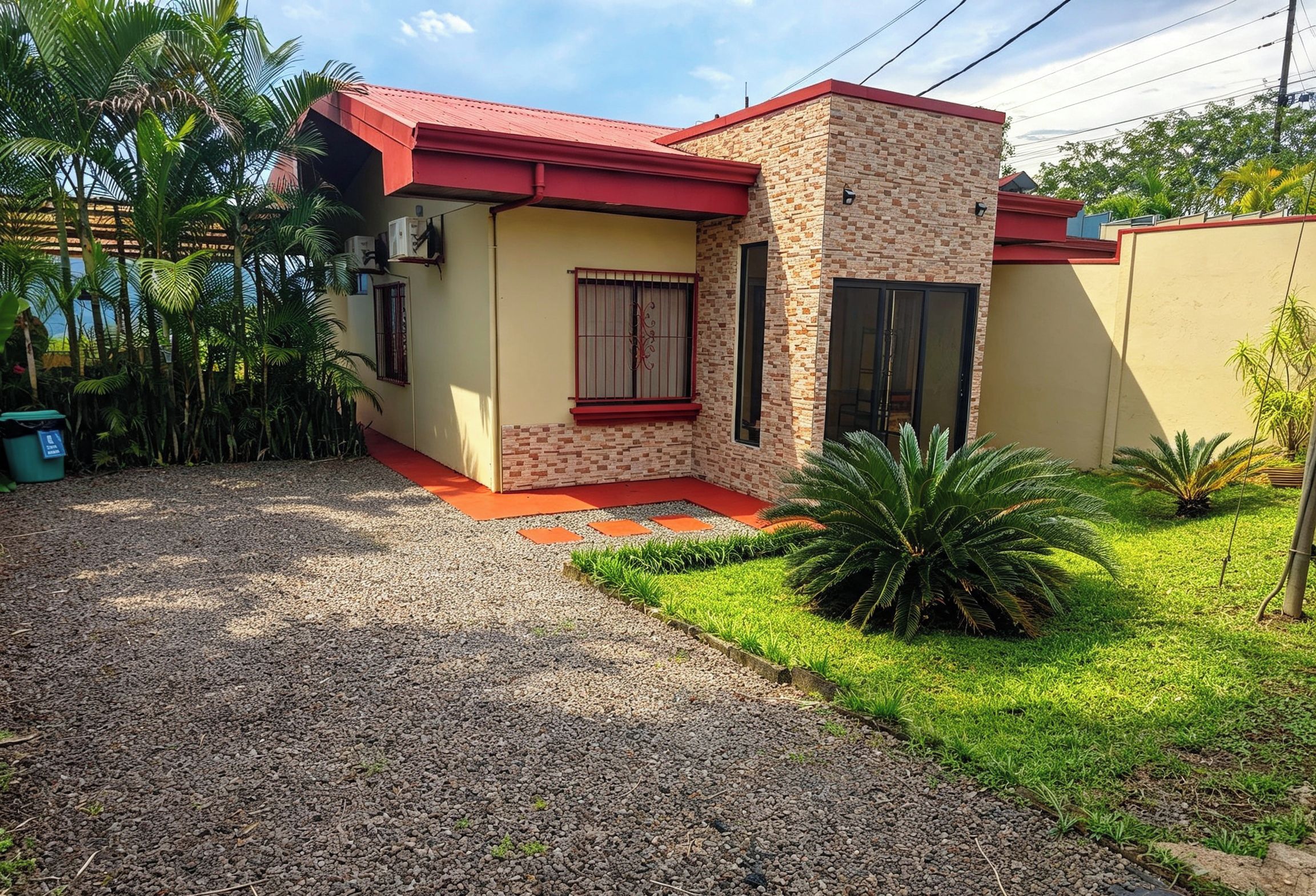 Un bâtiment à deux étages, avec un toit rouge, une façade en briques et un petit jardin devant, agrémenté d’un chemin de gravier et de plantes tropicales.