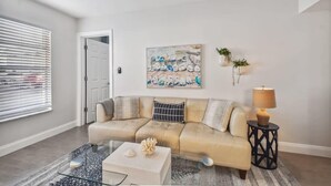Uma sala de estar com um sofá bege, uma mesa de centro de vidro, um quadro na parede e uma porta.