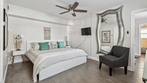 Quarto com cama, poltrona, ventilador de teto, televisão e um espelho grande.