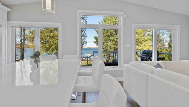 A modern interior with a white dining area, white sofas, and large windows offering views of a lake and trees. - Modern Waterfront Home on Georgian Bay w/ Hot Tub! (Victoria Harbour)