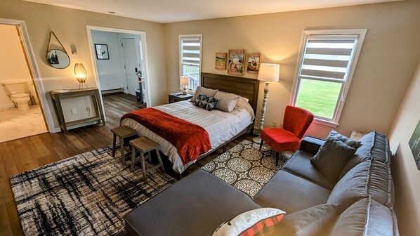 Une chambre spacieuse avec un grand lit, un fauteuil rouge, un canapé gris et un tapis à motifs.