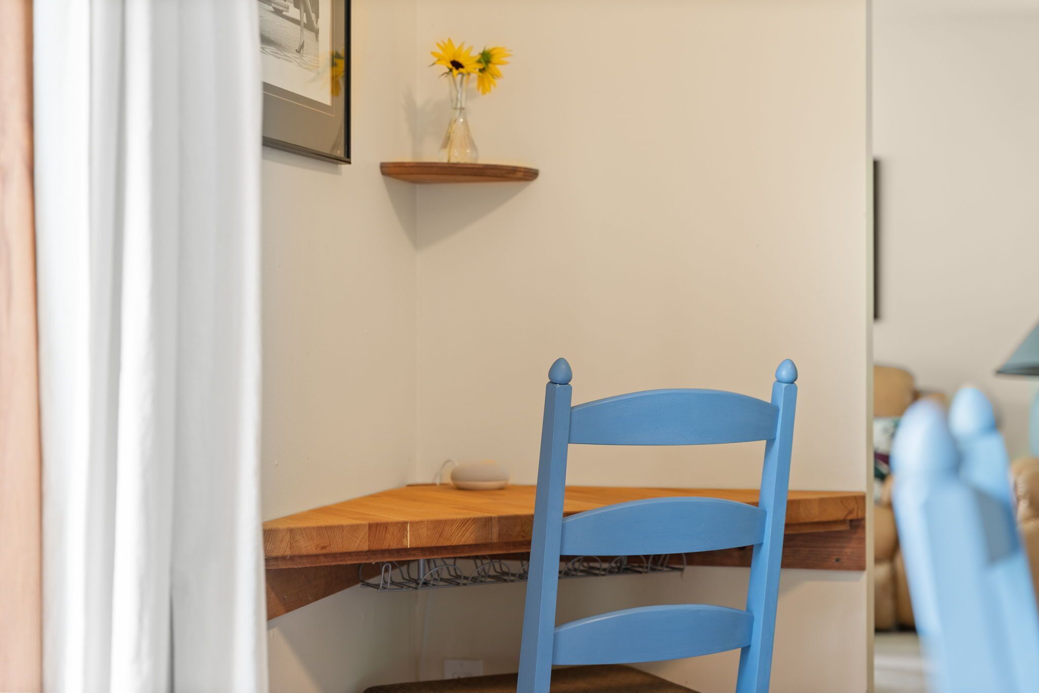 Un coin repas avec une table en bois, une chaise bleue et un vase rempli de fleurs jaunes, placé sur une étagère en hauteur.