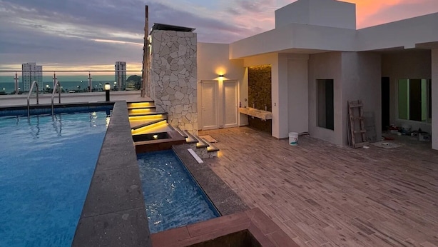Un área de piscina en la azotea con pared de piedra, cubierta de madera y vista al horizonte de la ciudad al atardecer.