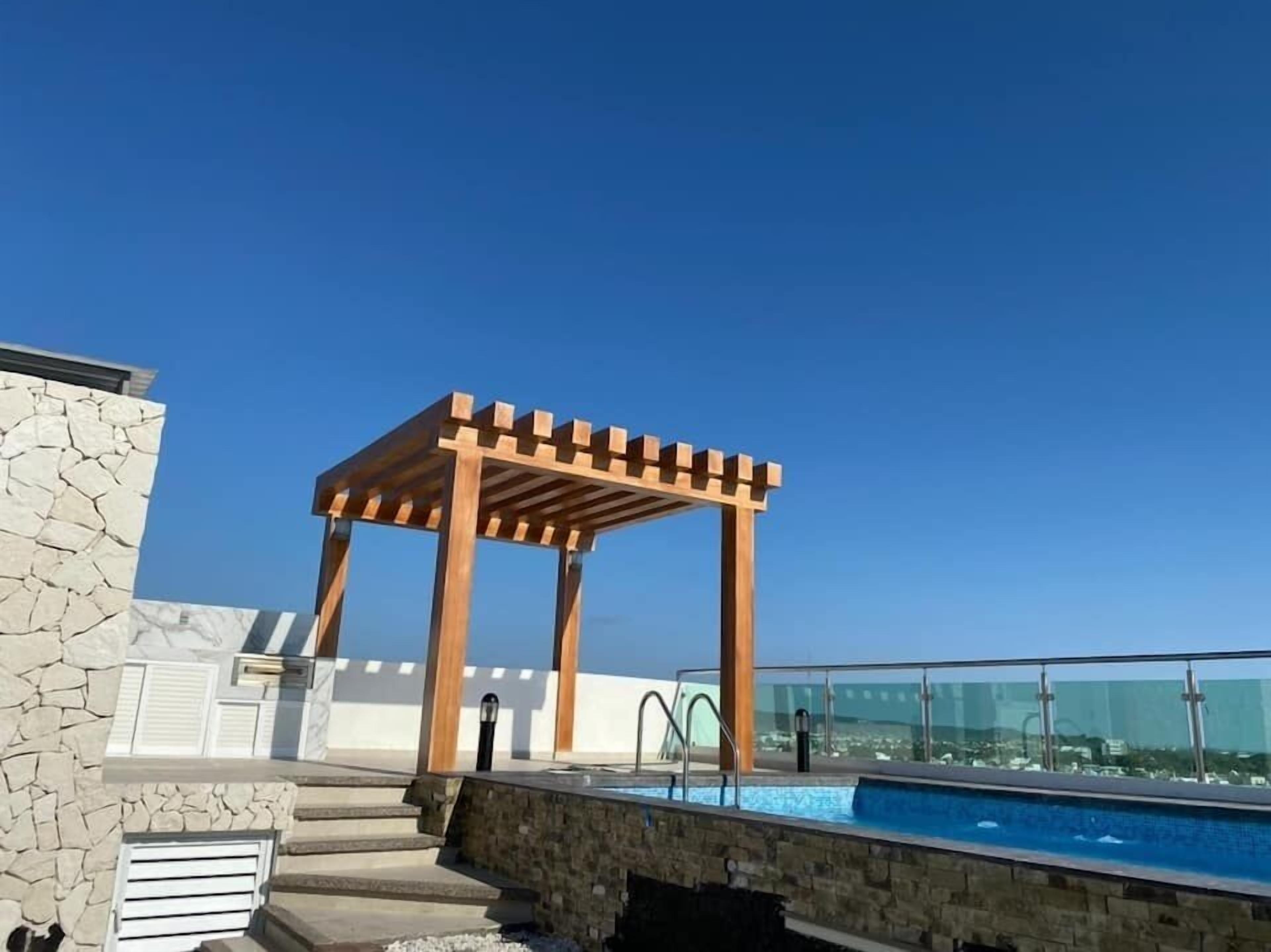 A rooftop pool area with a wooden pergola, stone steps, and a glass balustrade overlooking a cityscape.