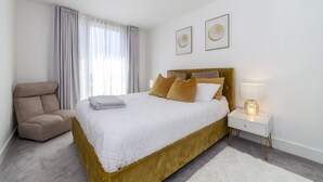 Un dormitorio moderno con una cama grande, una mesita de noche con lámpara, una silla y dos cuadros en la pared.