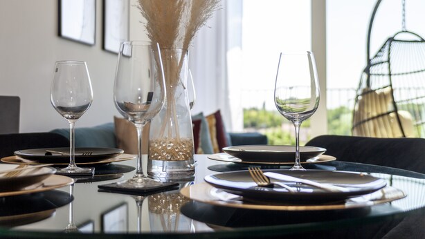 Una mesa de comedor dispuesta con platos, cubiertos y copas de vino, con un jarrón de plumas de plumas en el fondo.