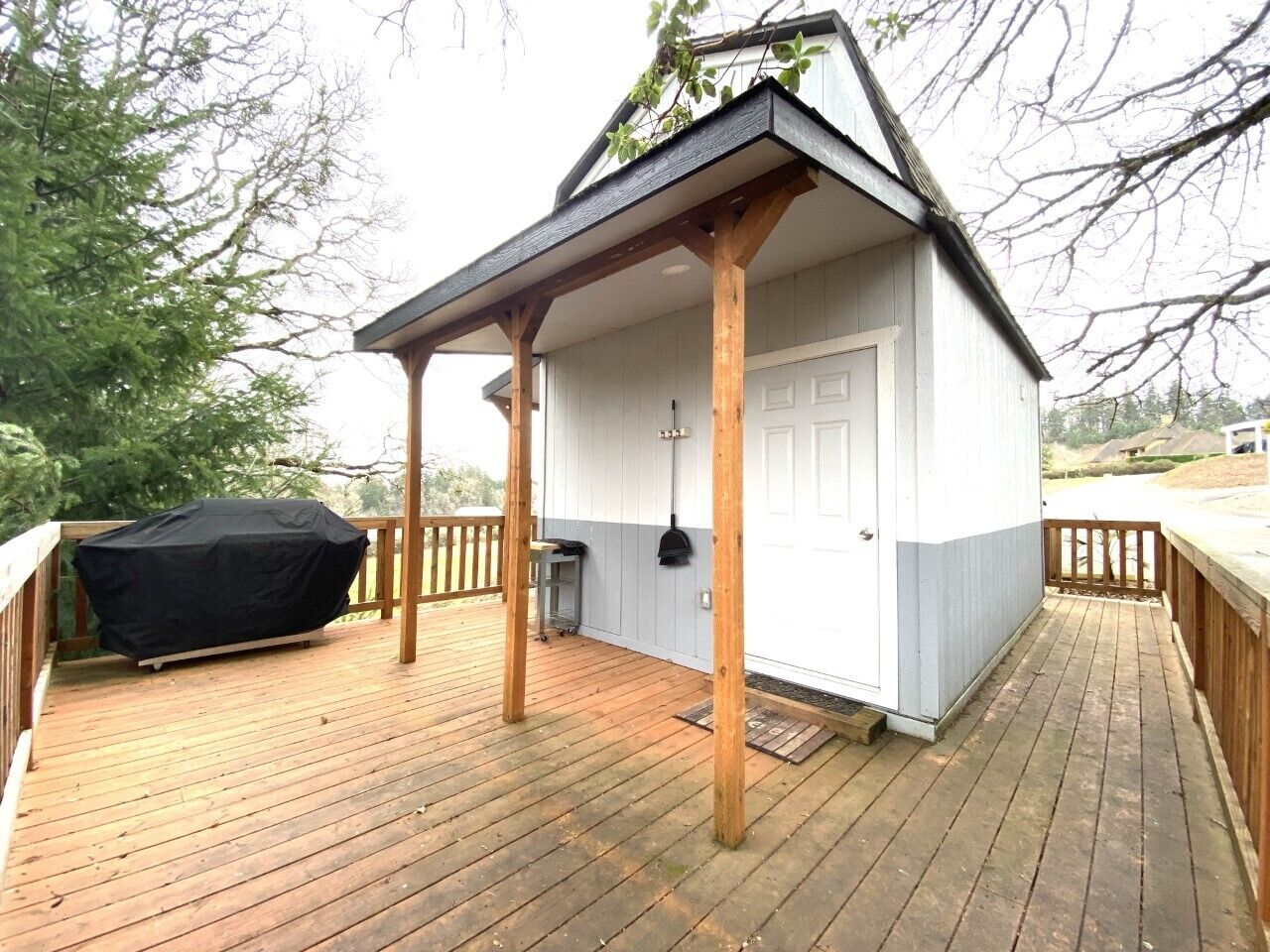 A covered outdoor area with a wooden deck, a barbecue grill, and a small building with a door.