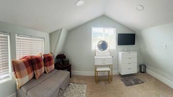 Un dormitorio con techo inclinado, una cama con manta a cuadros, un escritorio con silla, un televisor y una ventana con persianas.