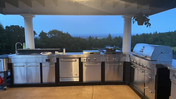 Cucina esterna con elettrodomestici in acciaio inossidabile, tra cui una griglia, situata in una zona coperta con vista su alberi e colline.