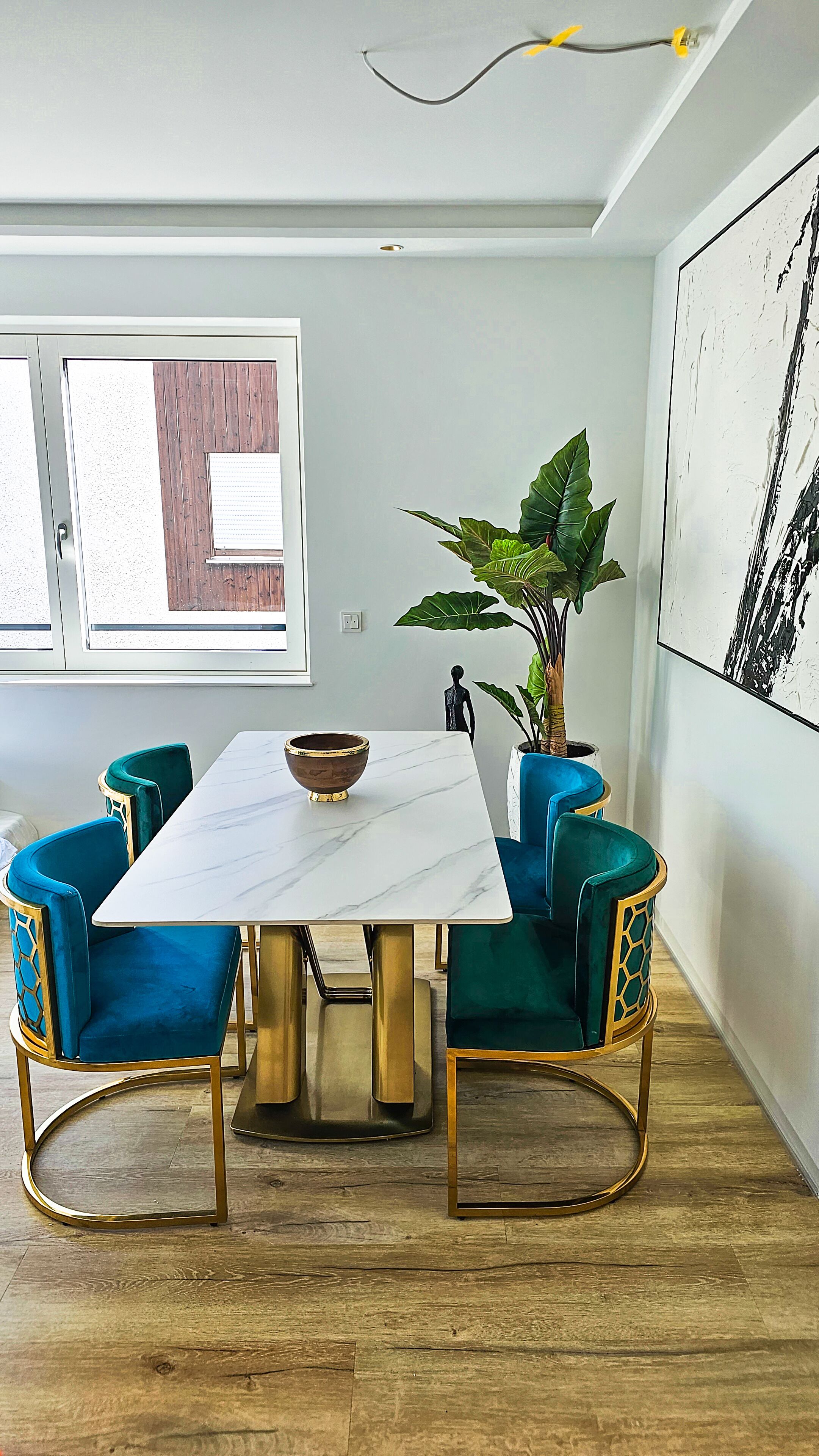 Uma sala de jantar com uma mesa de mármore, cadeiras estofadas em azul, um vaso com planta e um quadro abstrato grande na parede.