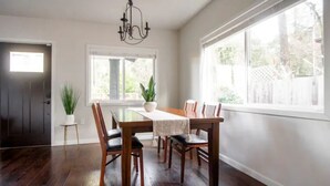 A dining area with a wooden table and chairs, a chandelier, and large windows with a view of greenery outside.