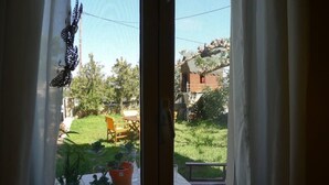 En vy genom ett fönster med en gardin, som visar en trädgård, en trästol och ett bord.