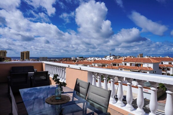 A rooftop terrace with a table and chairs, a view of the cityscape, and a white balustrade.
