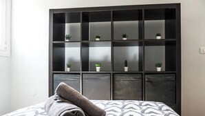 Un lit bien fait, recouvert d’une couvre-lit à motifs, et une serviette enroulée posée sur le bord. Au fond, on aperçoit une armoire en bois sombre, dotée de plusieurs étagères, dont certaines supportent de petites plantes en pot.