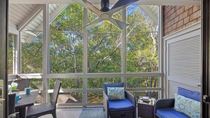 A covered porch with wicker furniture, a ceiling fan, and large windows offering a view of trees.