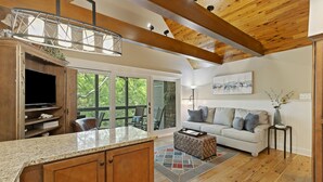 A spacious living area with a kitchen, wooden ceiling beams, a large window, and a cozy seating area.