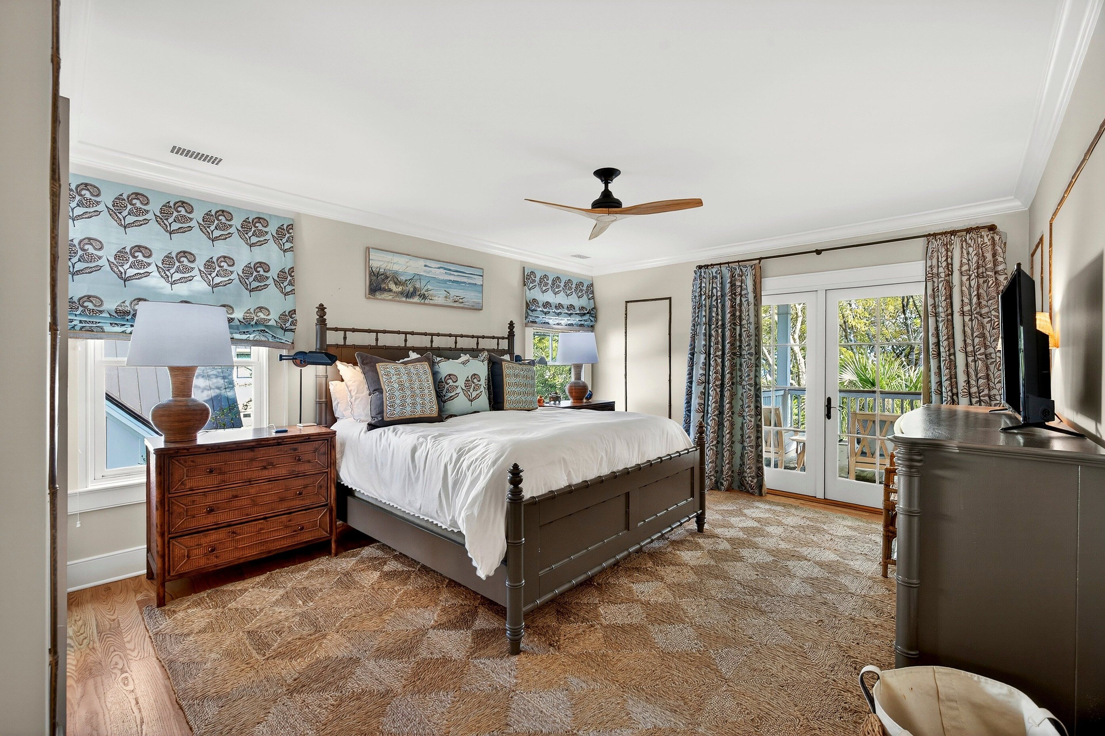 Une chambre à coucher comprenant un lit, des tables de chevet, une commode, un ventilateur de plafond et une vue sur l’extérieur, visible à travers une porte vitrée.