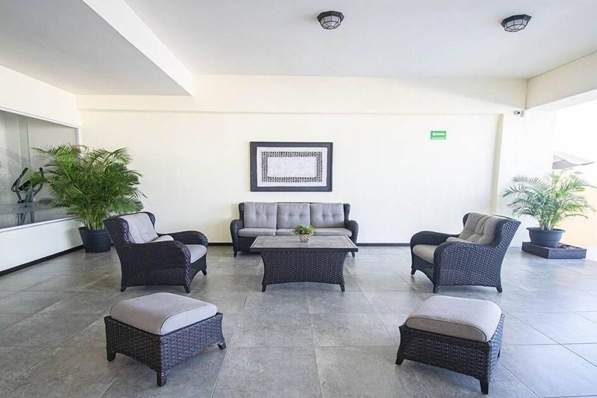 A spacious lobby with a grey sofa, two armchairs, and a coffee table. There are potted plants and a framed artwork on the wall.