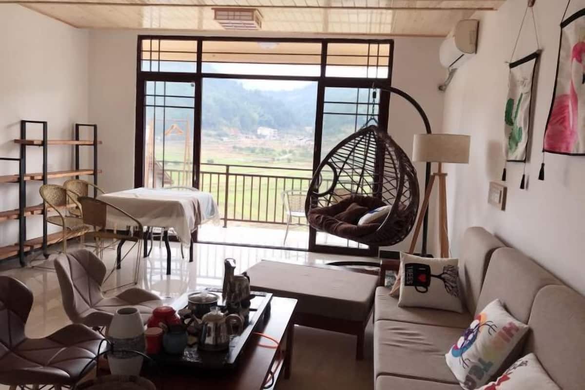 Een kamer met een bank, stoelen, een hangstoel, een tafel met dienblad en uitzicht op een landschap door grote ramen.