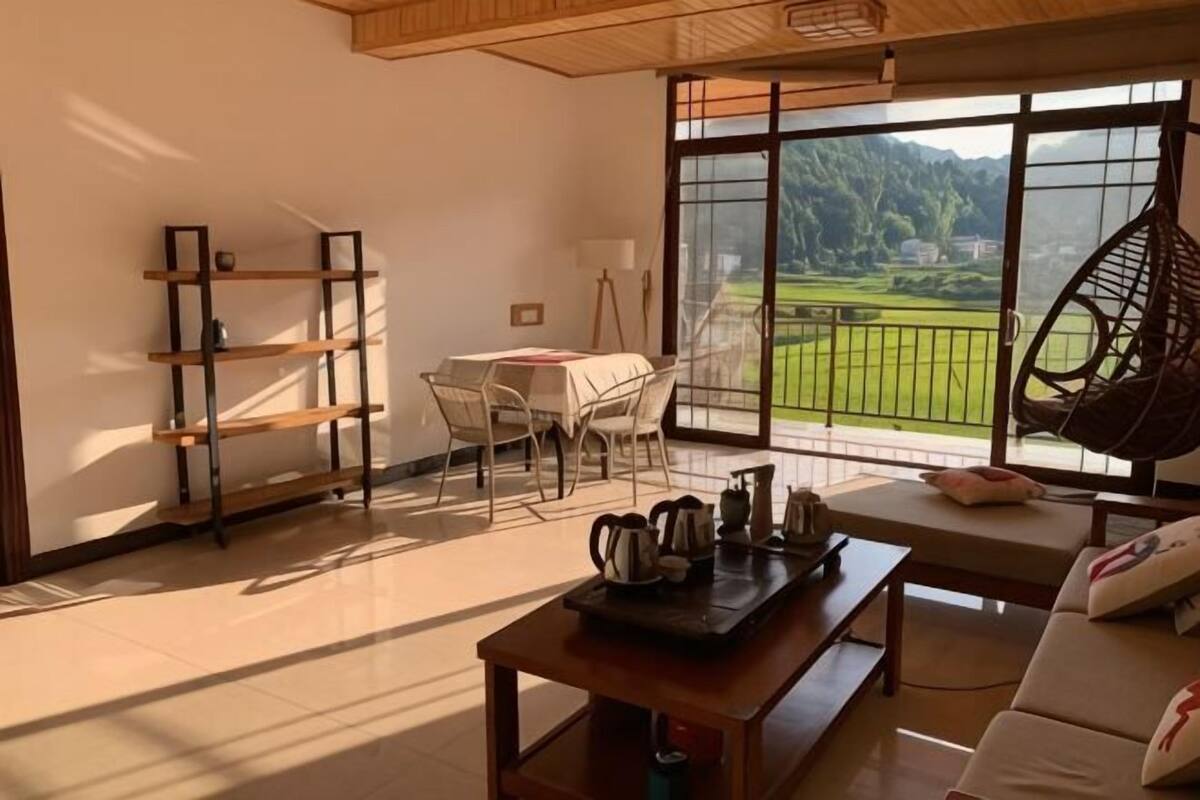 Een woonkamer met uitzicht op een groen landschap, een houten tafel met een waterkoker, een bank en een stoel.