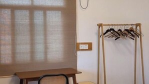 Ein Zimmer mit Holztisch, Stuhl, Wäscheständer und einem Fenster mit durchsichtigem Vorhang.