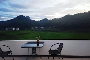 A balcony with a table and two chairs, overlooking a green landscape with mountains and buildings.