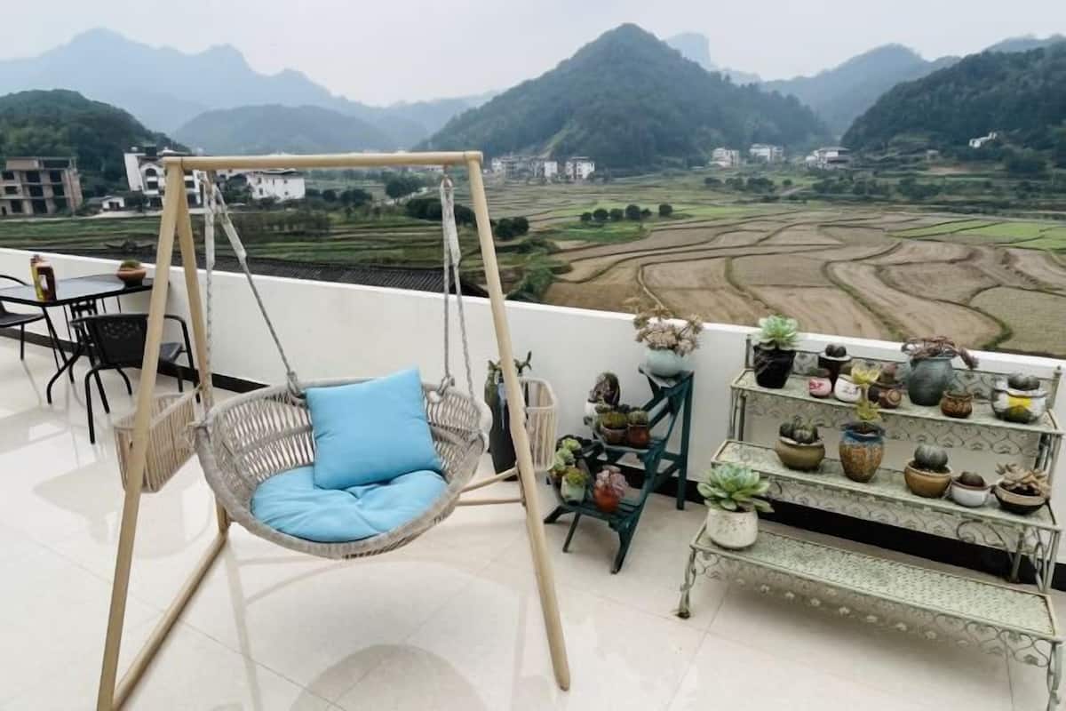 Een balkon met een rotan schommelstoel, een eettafel en stoelen. Er staan potplanten op een plank en achtergrond is er een uitzicht op terrassen en bergen.