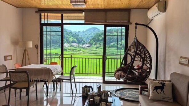 Ein Zimmer mit Esstisch, Stühlen, einem Hängesessel, einer Couch und Blick auf eine grüne Landschaft durch große Fenster.