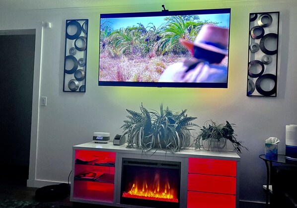 A living room with a flat-screen TV mounted on the wall, a fireplace with a red-lit glass front, and decorative wall art.