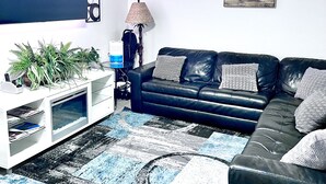 Een woonkamer met een zwarte leren bank, een witte tv-meubel met een aquarium, een vloerkleed met dessin en een schilderij aan de muur.