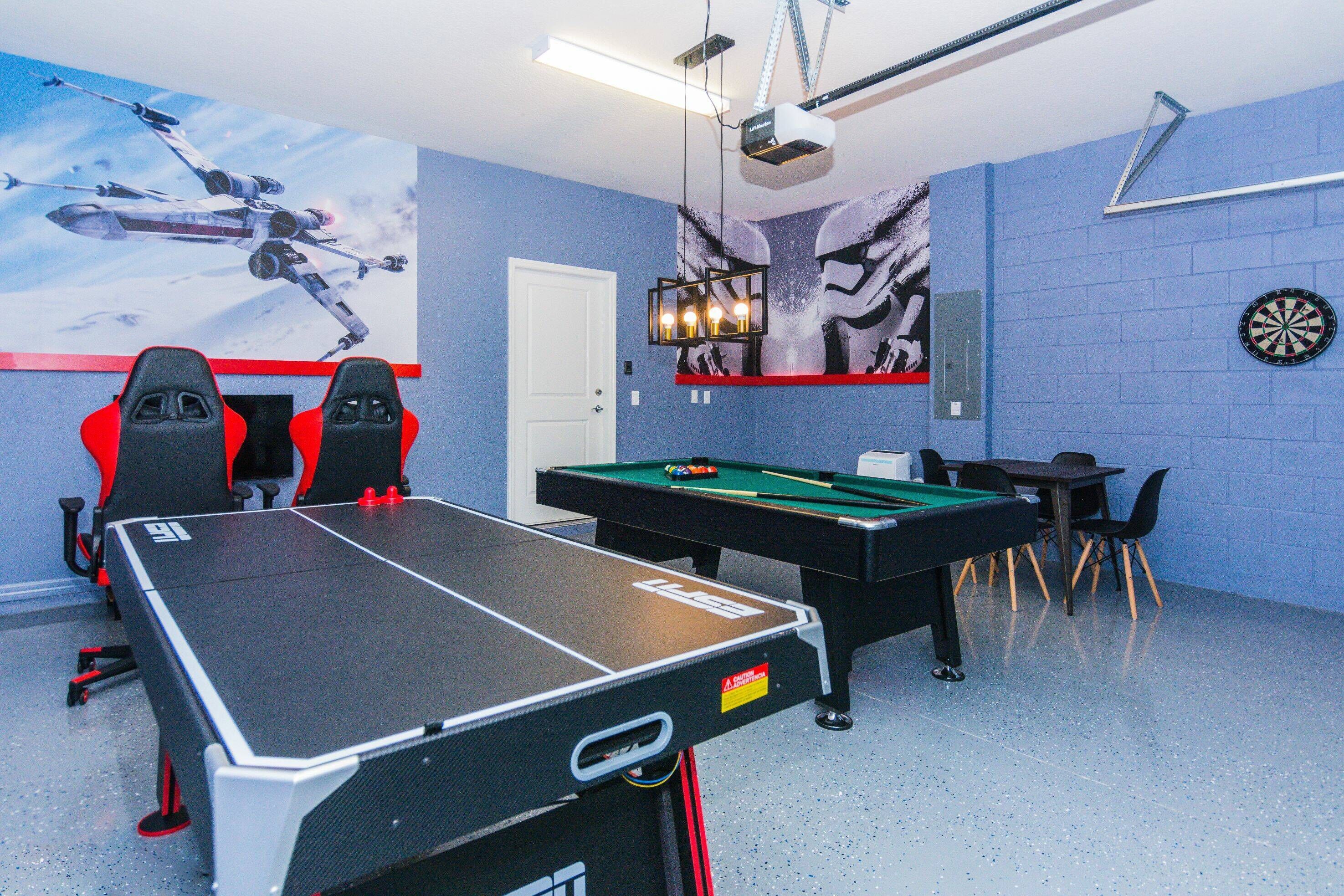Ein Raum mit Billardtisch, zwei roten Gaming-Stühlen, Dartscheibe und einer an der Decke befestigten Projektorleinwand.