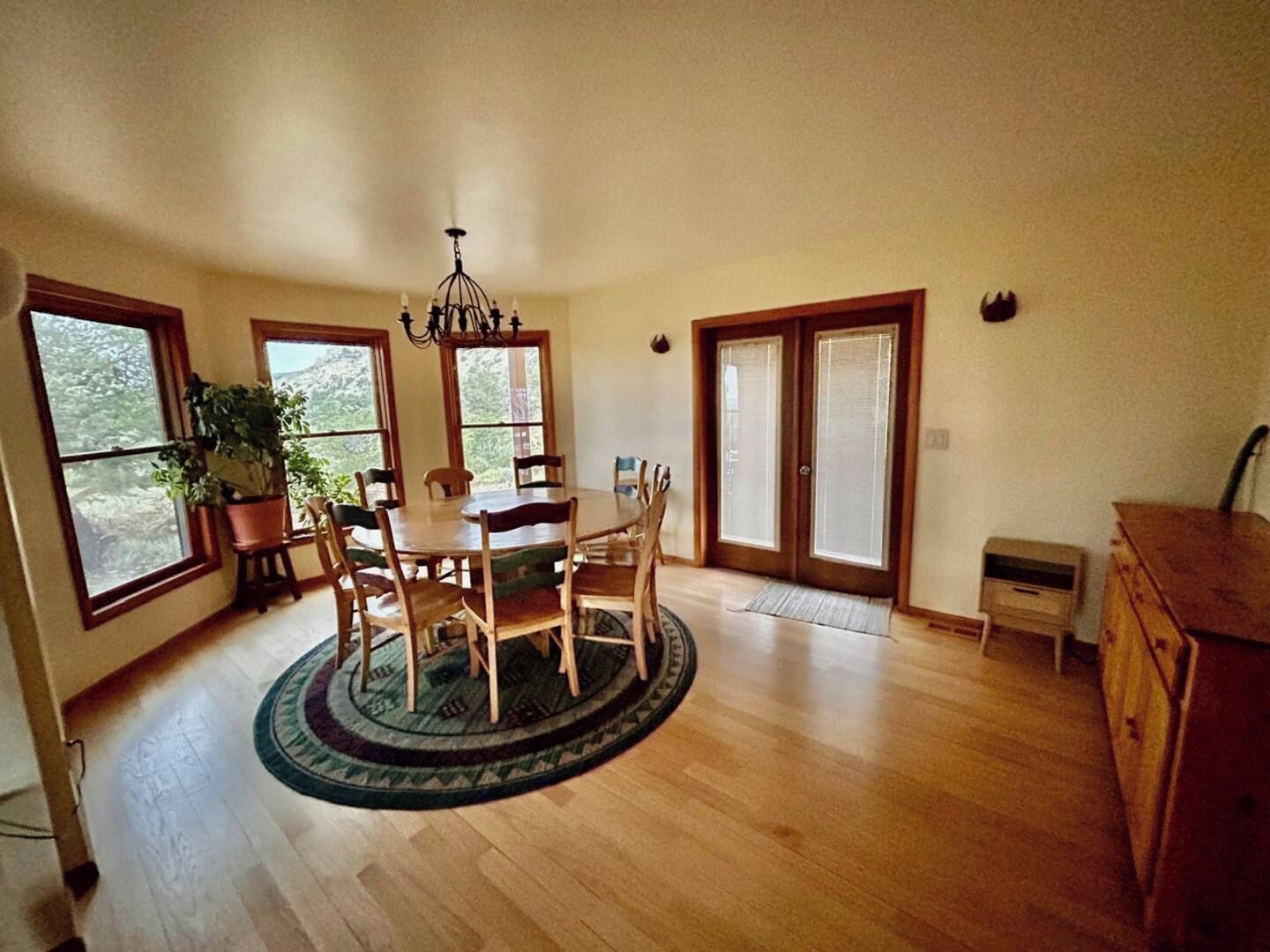 A dining area with a round table, chairs, a chandelier, large windows, and a wooden cabinet.