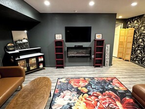 A living room with a flat-screen TV mounted above a fireplace, a sofa, a coffee table with a floral rug, and a bookshelf.