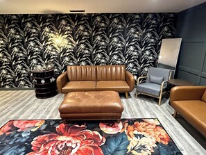 Uma sala de estar moderna com um sofá de couro marrom, um banquinho combinando e uma poltrona cinza. O ambiente possui um tapete estampado com motivos florais e uma parede com papel de parede de folhas tropicais.