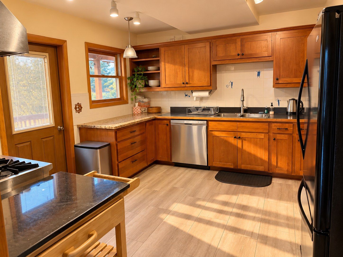 A kitchen with wooden cabinets, a stainless steel dishwasher, a black refrigerator, and a window with blinds.