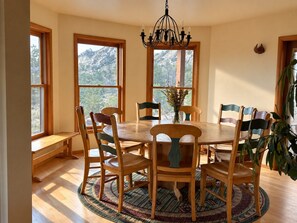 Ein Esszimmer mit einem runden Tisch, Holzstühlen, einer Kristallleuchter und großen Fenstern mit Blick auf die Berge.