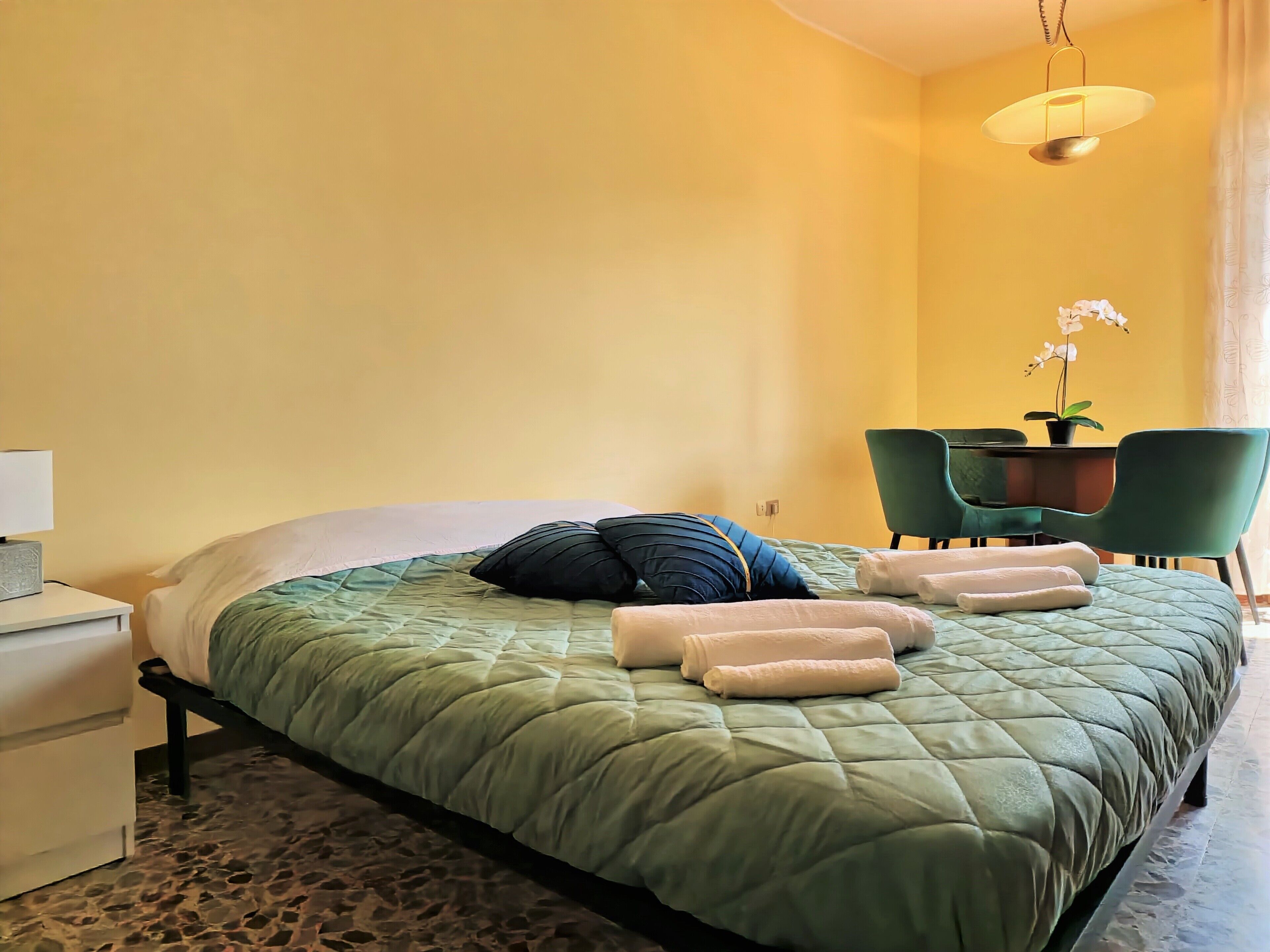 A neatly made bed with a green quilt, two pillows, and rolled towels on a stone-patterned floor.