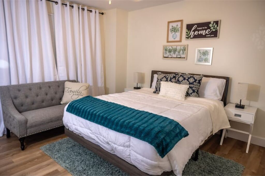 A bedroom with a bed, a grey sofa, a bedside table, and a wall with framed pictures.