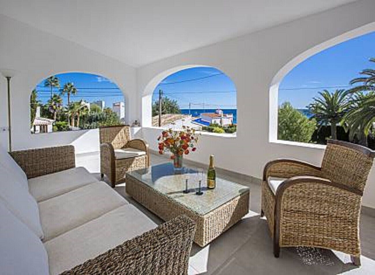 A sunlit balcony with wicker furniture, a glass table with a bottle, and a view of the sea and buildings.