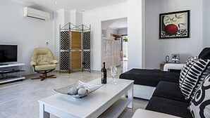 Een moderne woonkamer met een witte bank, een kussen met zwart-wit patroon, een witte salontafel met decoratieve objecten, een beige fauteuil en een tv-meubel.