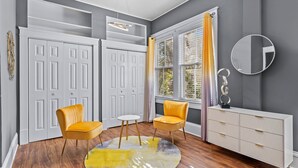 A room with a white wardrobe, two yellow chairs, a small white table, a round rug, a white console table, and a mirror on the wall.
