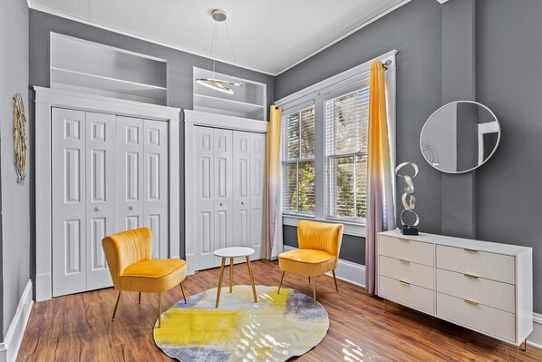 A room with a white wardrobe, two yellow chairs, a small white table, a round rug, a white console table, and a mirror on the wall.