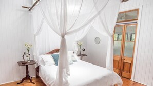 Un dormitorio con cama con dosel, paredes blancas, suelo de madera, una mesita de noche pequeña con lámpara y una puerta de madera.