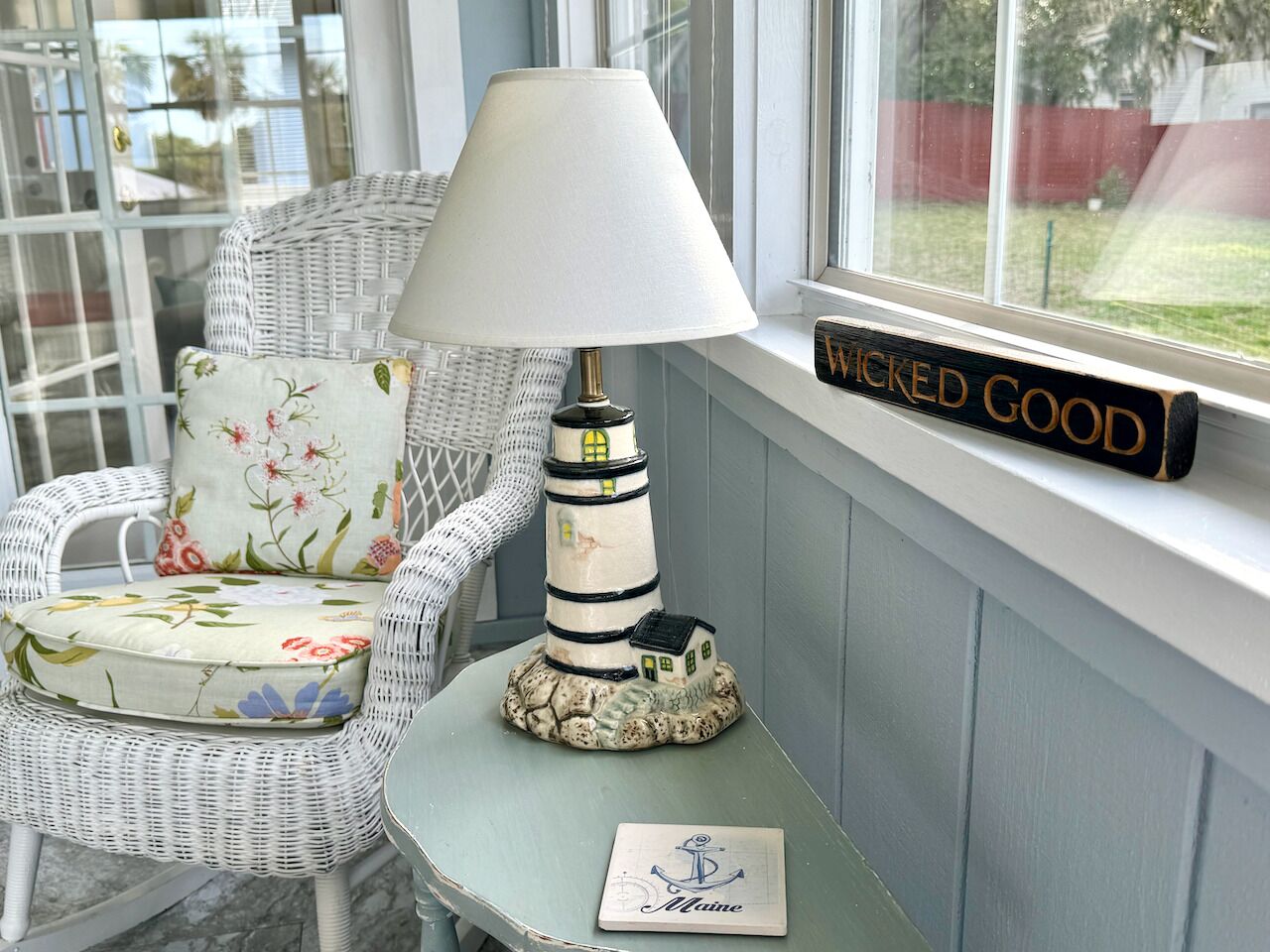 A wicker chair with floral upholstery, a table lamp, and a sign reading "WICKED GOOD" on a window sill.