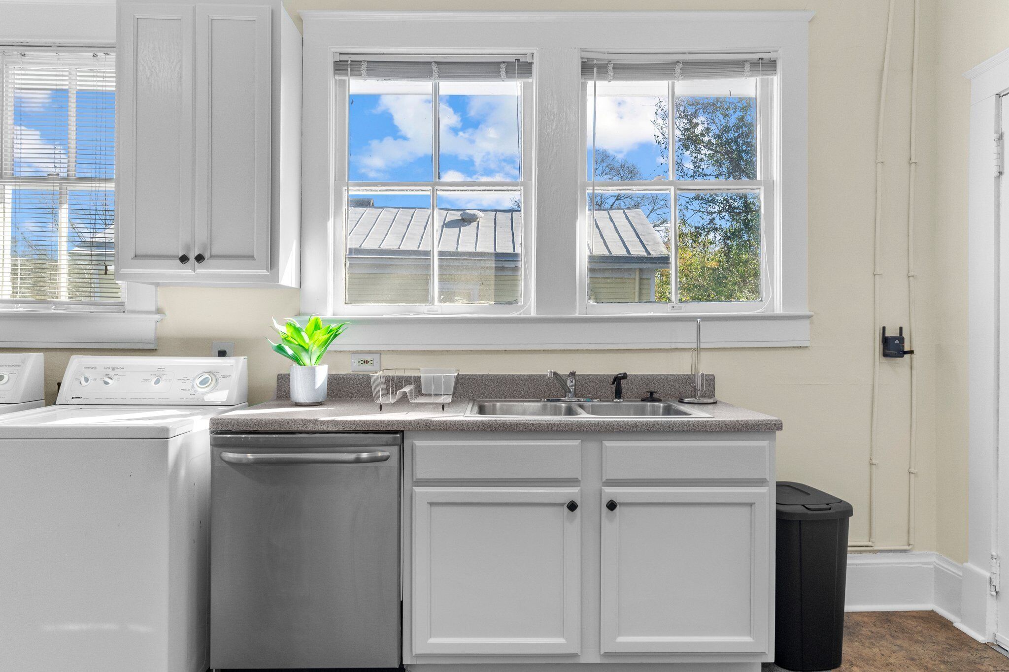 Una cocina con armarios blancos, lavavajillas, un lavabo y una ventana con vista a una casa y árboles.