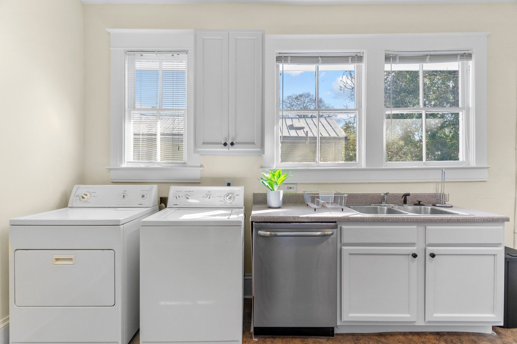 A laundry area with a washer, dryer, and dishwasher, white cabinets, and a window with blinds.